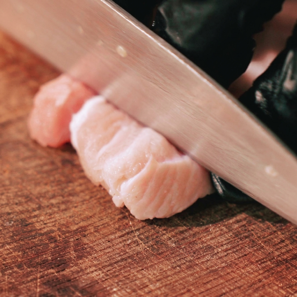 手袋をした手が、木製のまな板の上で生の皮付き豚バラ肉を太めの拍子木切りにしている様子。