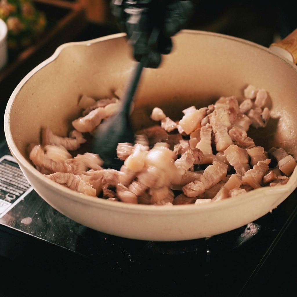 ベージュのフライパンで豚肉を黒いヘラで炒めている様子。焼き色がつき、脂が溶け出している。