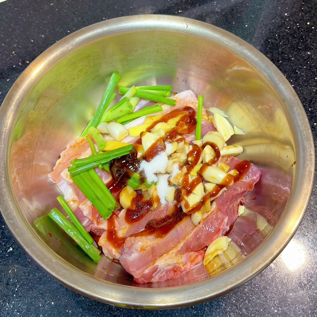 金属製ボウルの中で、マリネされた豚肉と香味野菜の上に、白い塩と赤茶色のスパイスパウダーが盛られている様子。