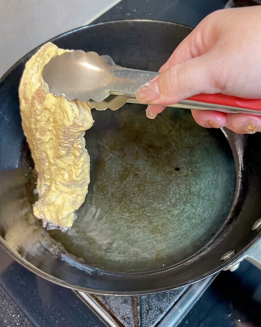 衣をまとった豚バラ肉を、金属製のトングを使ってフライパンの熱い油の中にそっと入れている様子。