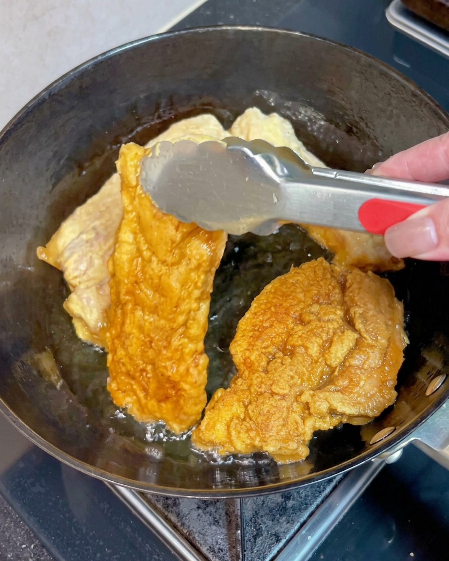 泡立つ熱い油の中で、他の肉片と一緒に、こんもりときつね色に揚がった豚バラ肉を金属製のトングで反転させている様子。