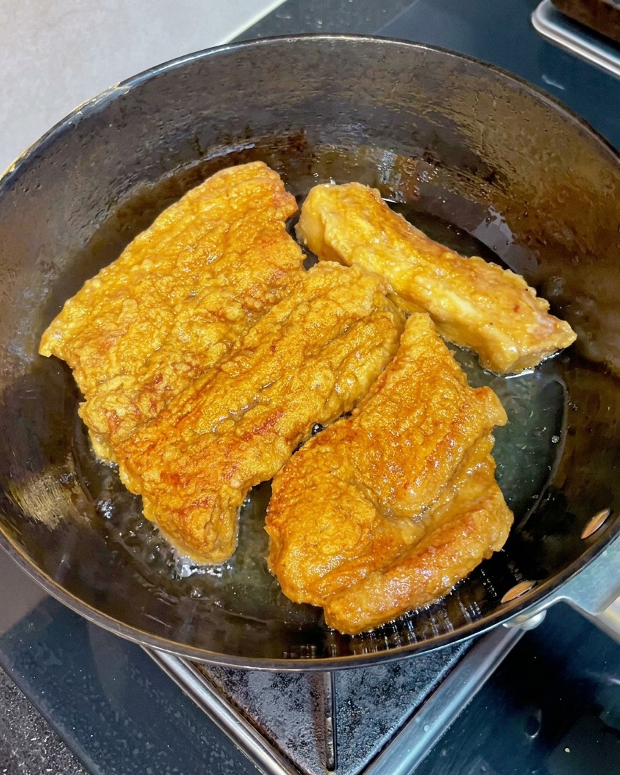 フライパンの熱い油の中で揚がっているカリカリのきつね色の豚バラ肉ブロックに、木製の箸を刺して柔らかさを確認している様子。