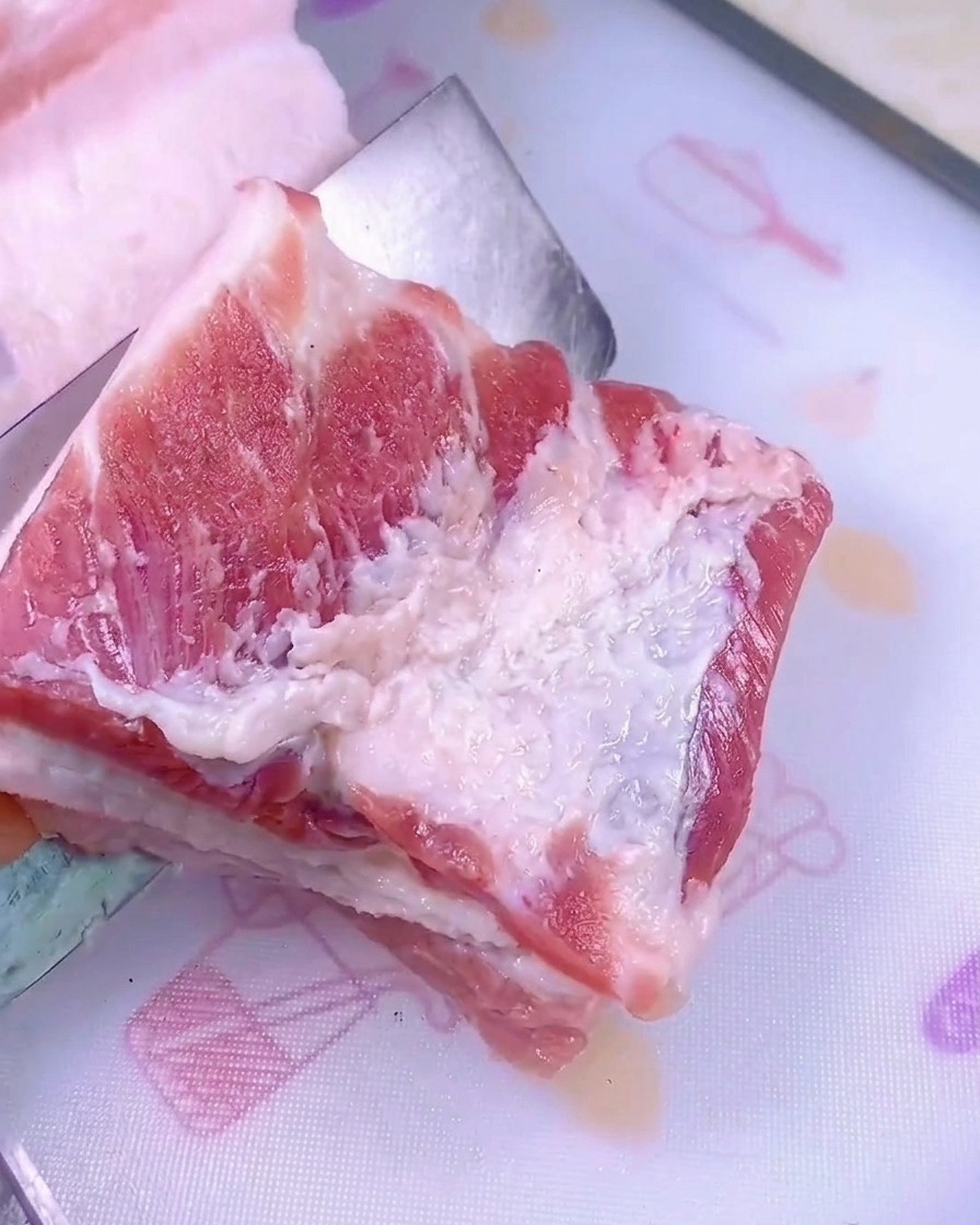 白いプラスチック製のまな板の上で、鋭い包丁を使って生の厚切り豚バラ肉をスライスしている様子。