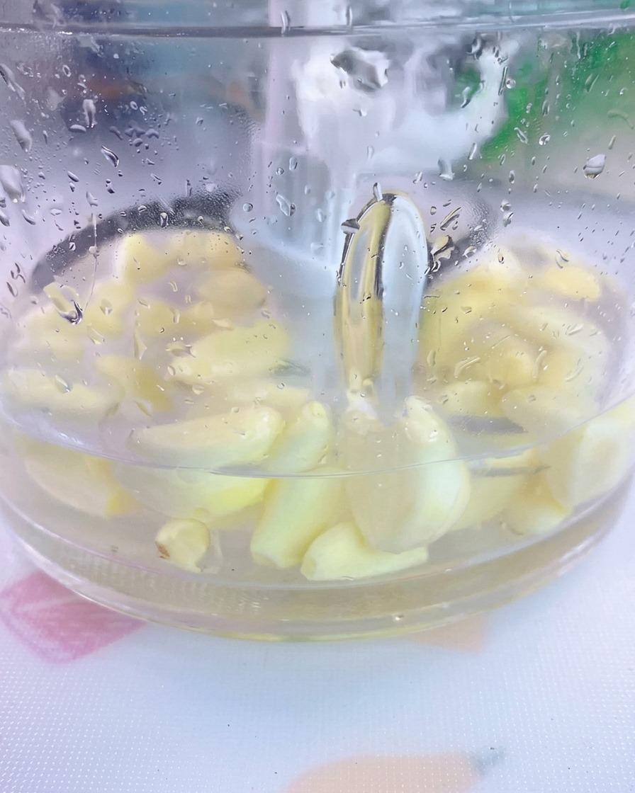 透明なミキサーの容器の中で、皮をむいたニンニクの芽と塩、水が混ざり合っている様子。