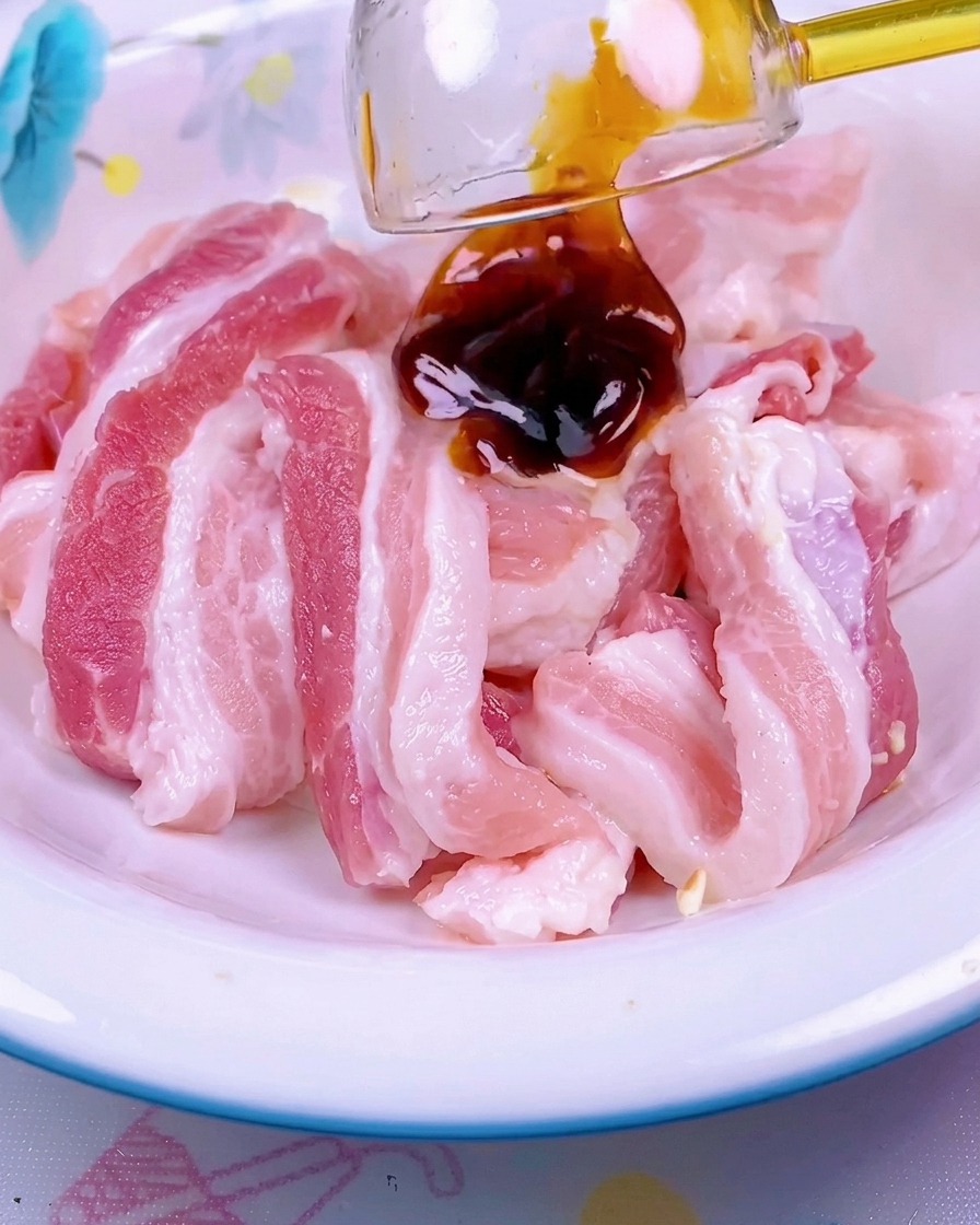 白いセラミック製のボウルに入ったスライスした生の豚バラ肉に、濃い赤色のソースが回しかけられている様子。