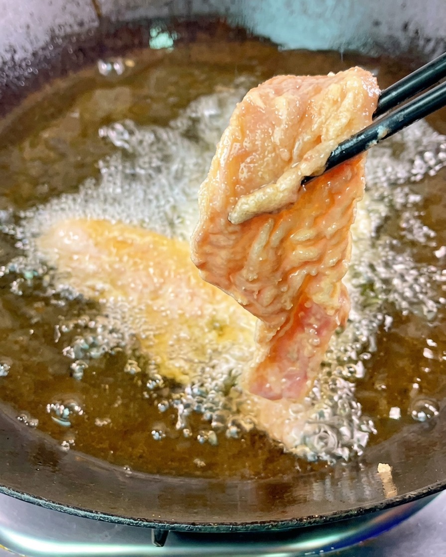 黒い箸を使い、衣をまとった豚バラ肉のスライスを1枚ずつ、ふつふつと波打つ熱い揚げ油に入れている様子。