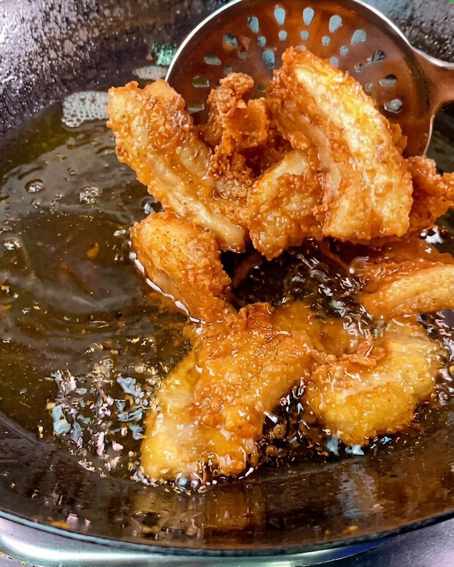 泡立つ中華鍋の油から、穴あきお玉を使って、こんがりと揚がったきつね色の豚バラ肉を引き上げている様子。