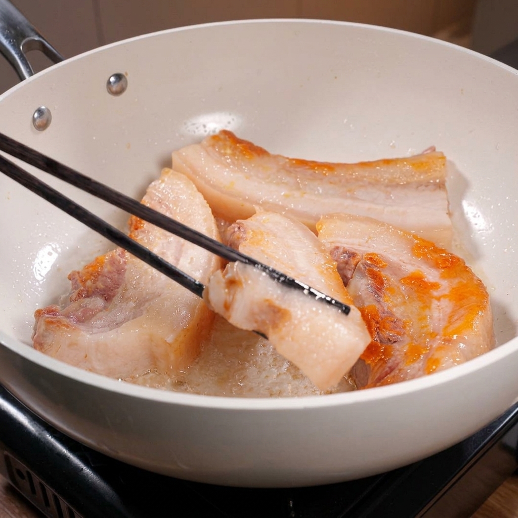 白いフライパンの中で、長い黒い箸を使って厚い豚バラ肉をひっくり返している様子。端のほうが美しく黄金色のきつね色に焼けているのが見える。