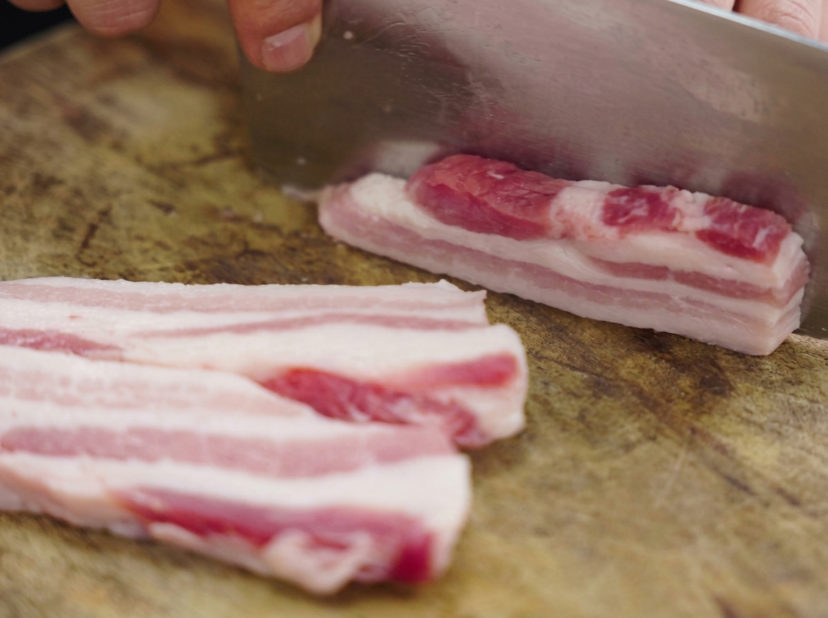 木製のまな板の上で、大きな中華包丁を使って生の豚バラ肉を厚めの短冊状に切っている様子。