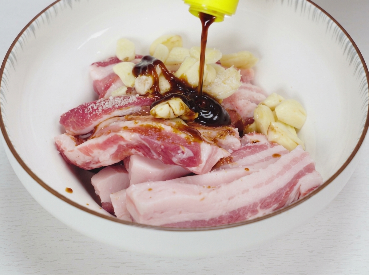 白い陶器のボウルに入った生の豚バラ肉と潰し大蒜の上から、濃い液体の調味料やオイスターソースを注いでいる様子。