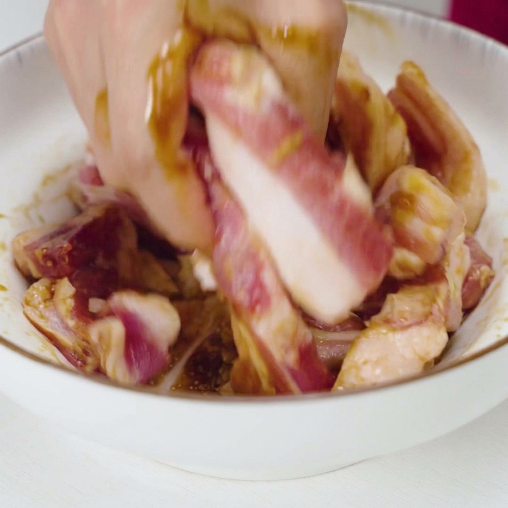 白い陶器のボウルの中で、手を使って生の豚バラ肉に濃い色の調味料と大蒜をしっかりと揉み込んでいる様子。