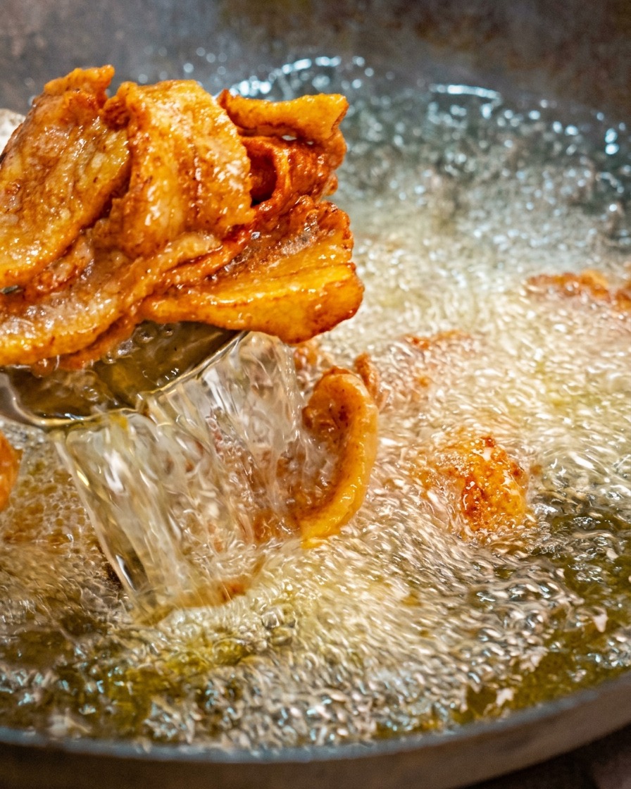 熱く泡立つ油の中華鍋から、網じゃくしを使って、きつね色に揚がって端が丸まった豚バラ肉のスライスをすくい上げている様子。