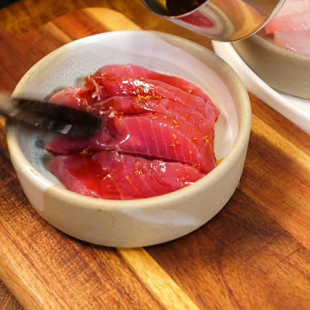 風情のあるセラミックの小鉢に入った鮮やかな赤身のマグロに、旨味たっぷりの生姜醤油タレをスプーンで回しかけている様子
