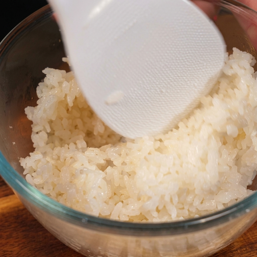 クリアなガラスボウルの中で、炊きたての白いご飯に合わせ調味料を加え、白いプラスチック製のしゃもじで優しく切るように混ぜている様子