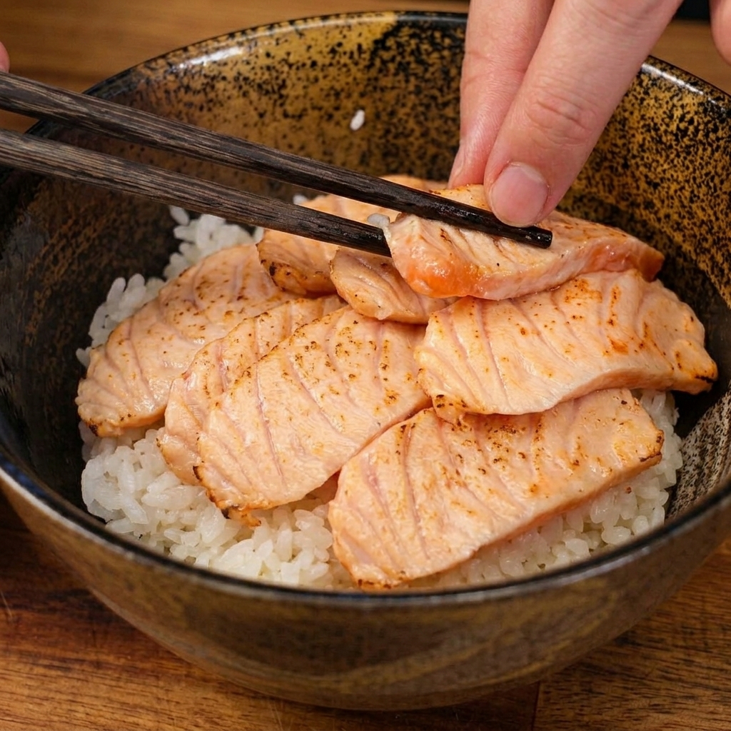 セラミックの丼に盛られた酢飯の上に、木製のお箸を使って、軽く炙ったサーモンの切り身を丁寧に配置している様子