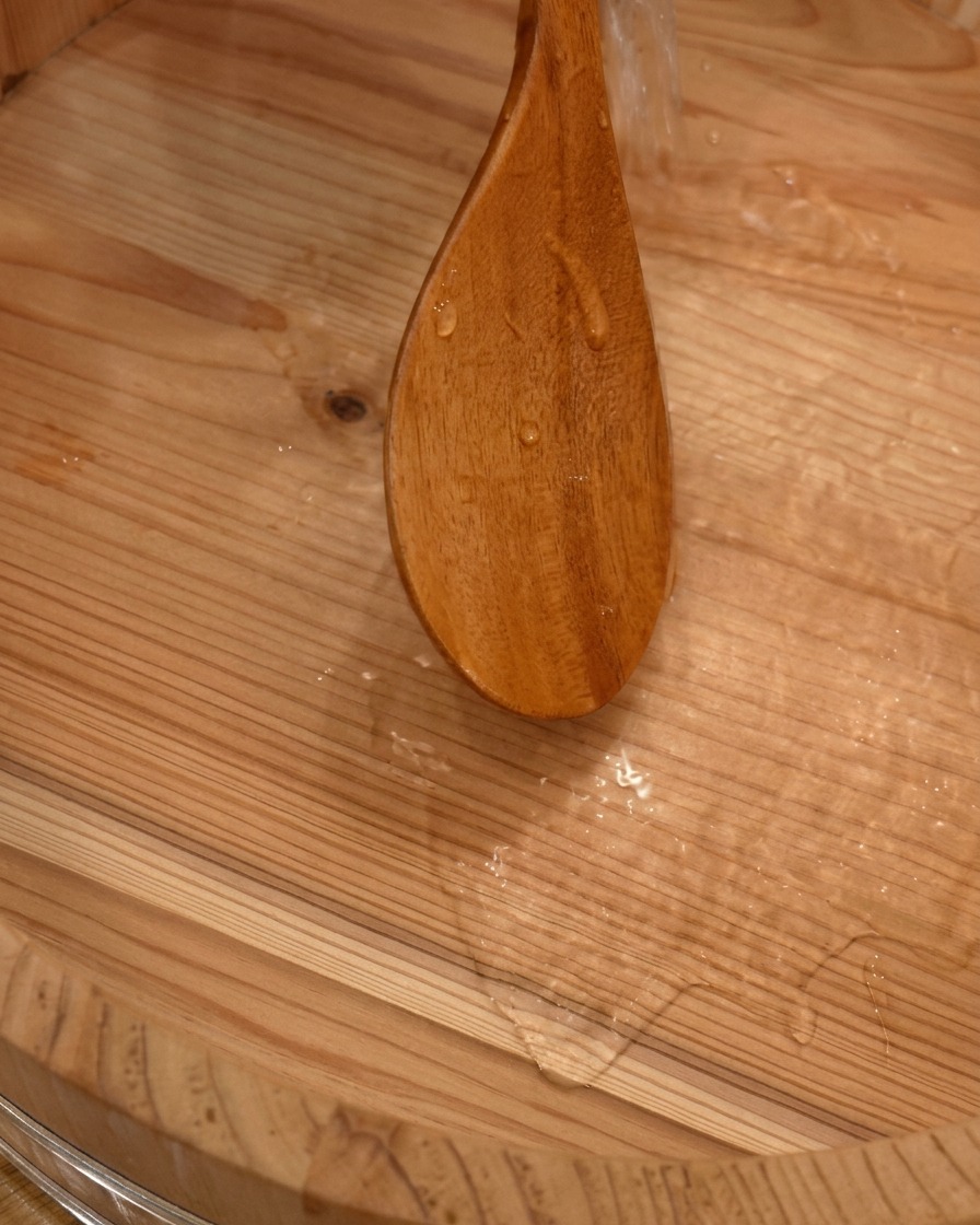 伝統的な大きな木製の寿司桶(飯台)に、しゃもじを使って水を注ぎ、内側を湿らせている様子。