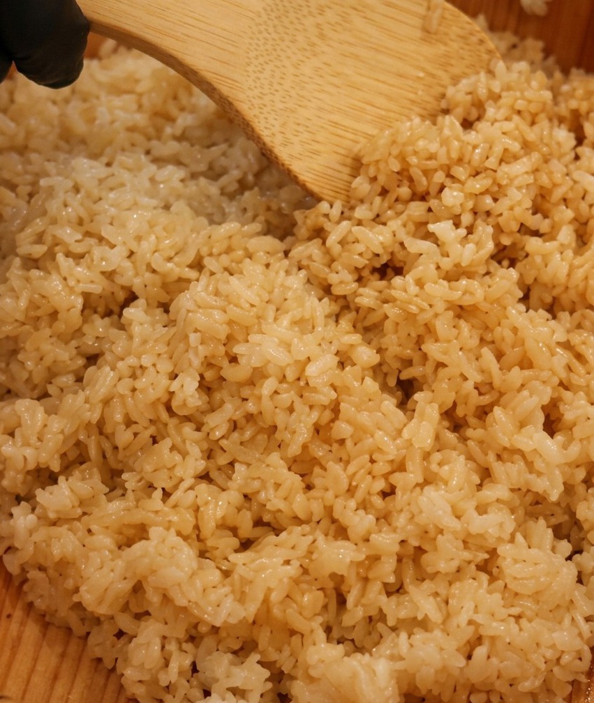 木製の寿司桶の中で、湯気の立つご飯にすし酢を回し掛け、木製のしゃもじで優しく混ぜ合わせている様子。
