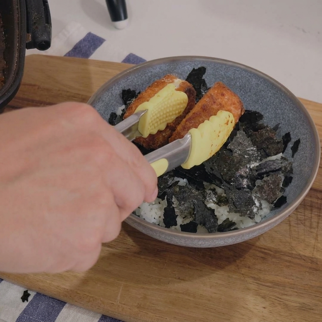 先端が黄色のトングを使い、きざみ海苔を敷いたグレーの丼のご飯の上に、こんがり焼けた鮭の蒲焼きを並べて盛り付ける様子。