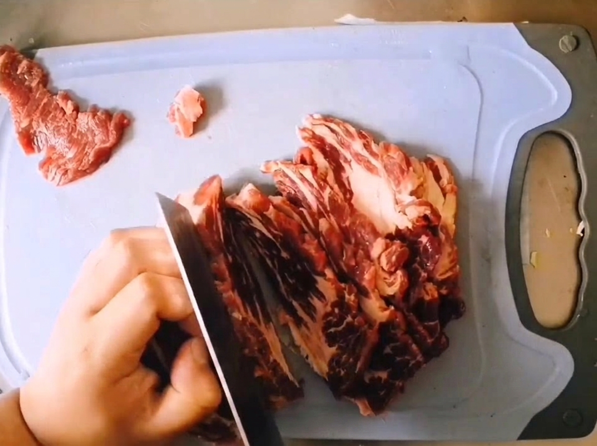 薄い青色のまな板の上で、よく切れる包丁を使って生の霜降り牛肩ロース肉を厚めに切り分けている様子。