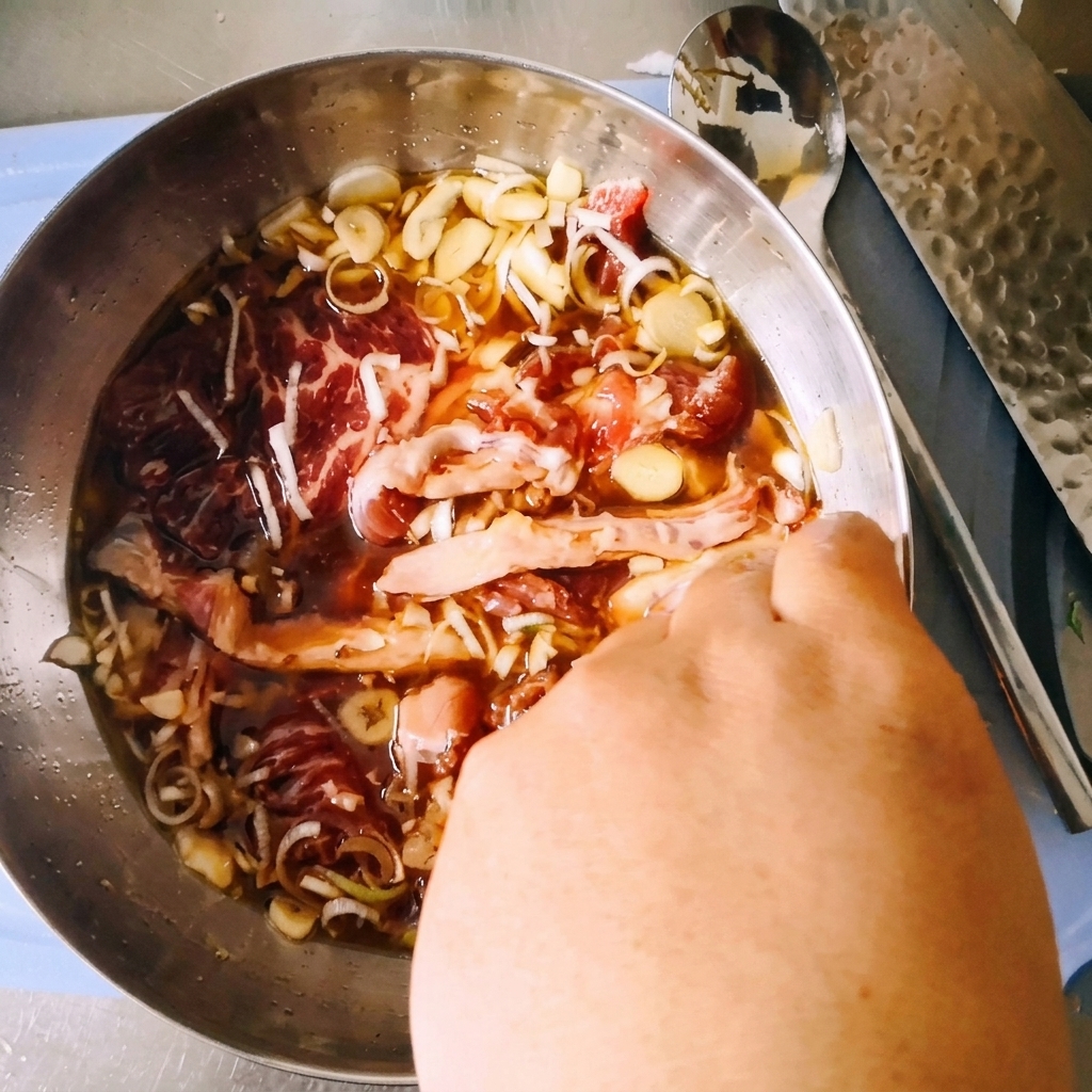 ステンレス製のボウルの中で、スライスした生の牛肩ロース肉を、濃い醤油ベースのタレやネギ、にんにくと一緒に手で揉み込んでいる様子。