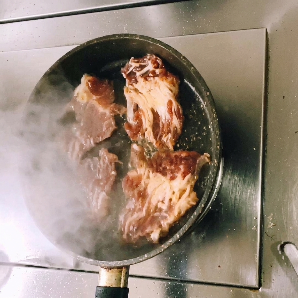 平らなフライパンでタレに漬けた牛肉のスライスを焼き、肉が茶色く色づき始めると同時に湯気が立ち上っている様子。