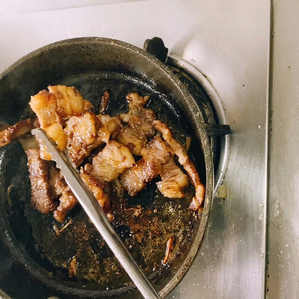 フライパンの上で、金属製のトングで焼き色のついた牛肉を持ち上げながら、キッチンバサミで小さく切り分けている様子。