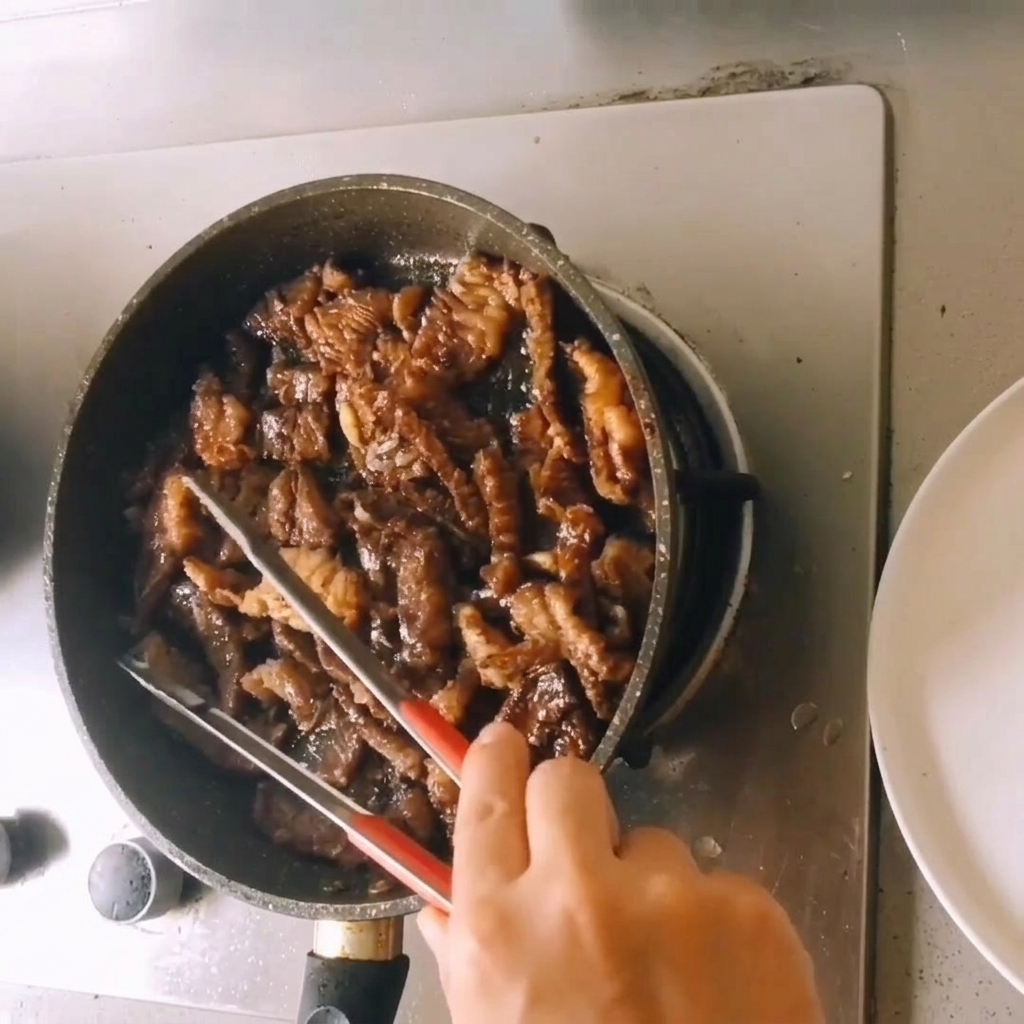 フライパンの中で、一口大に切った調理済みの牛肉をトングで和えながら、とろみのある濃い色のソースで煮詰めてツヤを出している様子。