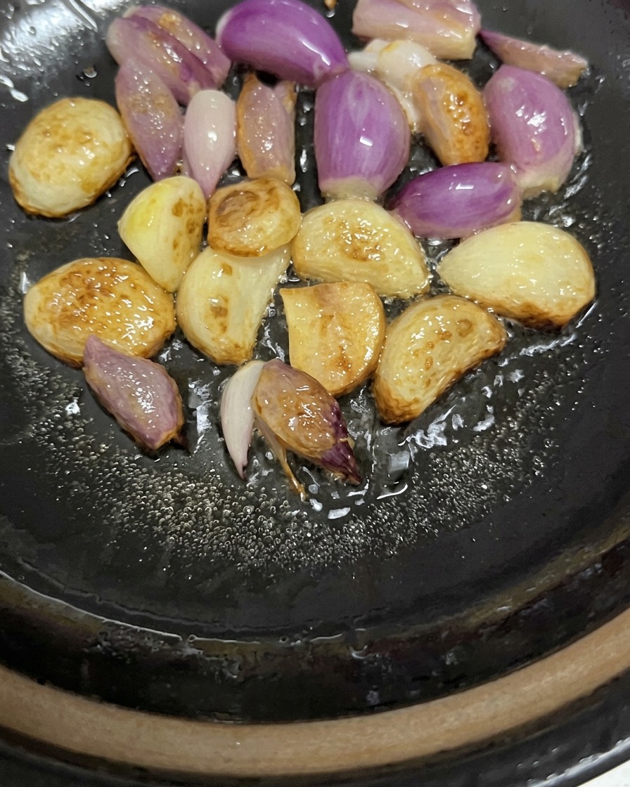 土鍋の底で、油をまといながらキツネ色に色づき、パチパチと音を立てて炒められる丸ごとのにんにくとエシャロット。