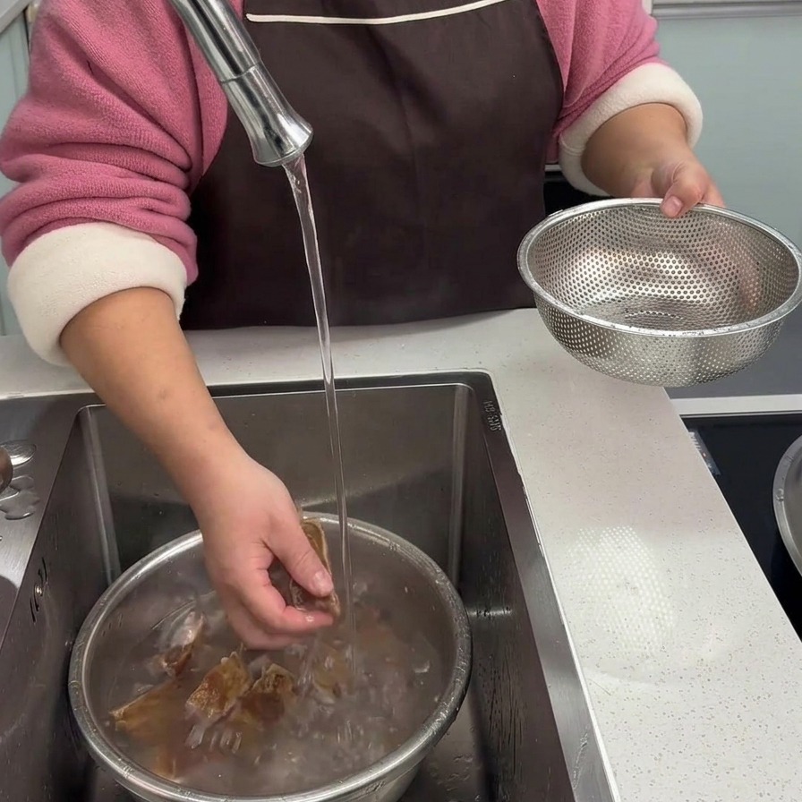 キッチンのシンクで、下茹でした干し豚スペアリブを冷たい流水で丁寧に洗っている手元。