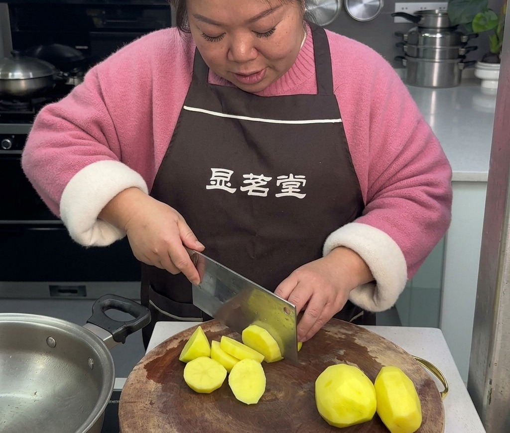 茶色のエプロンを着た女性が、丸い木製のまな板の上で皮をむいた黄色いジャガイモを大きな包丁で乱切りにしている様子。