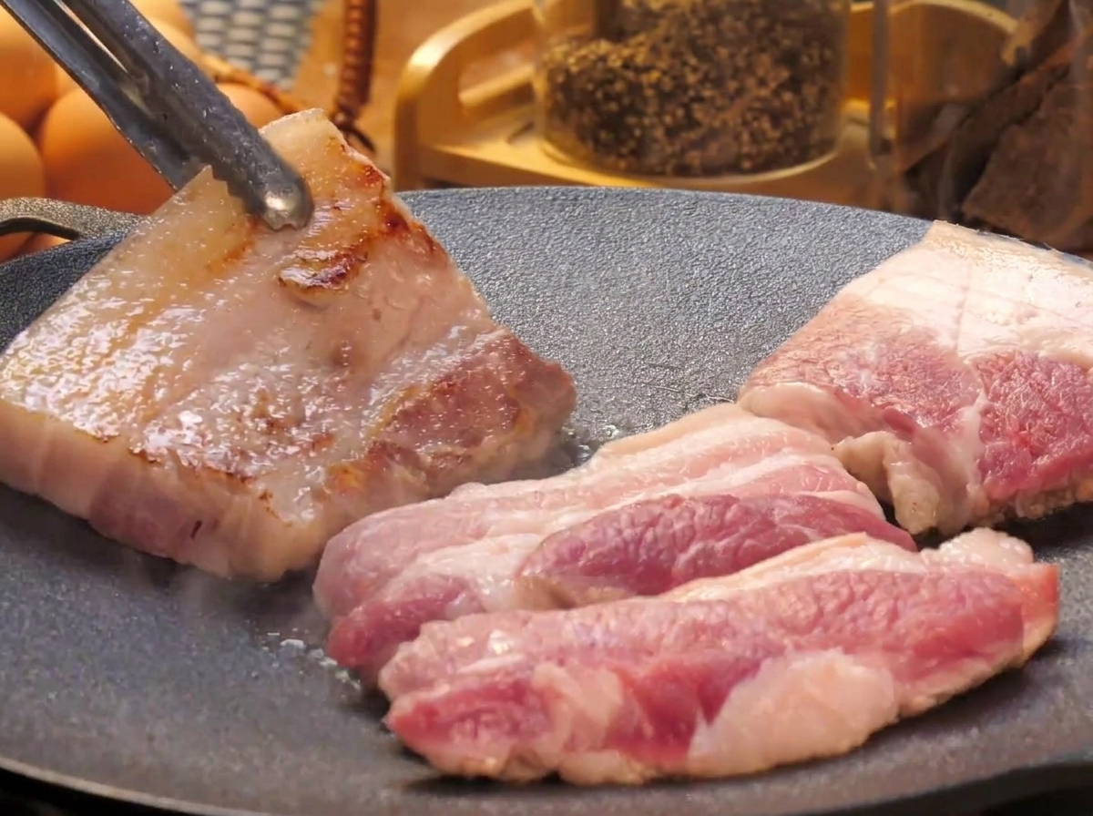 平らな鉄板の上で焼かれている厚切りの豚バラ肉を、トングを使って裏返し、こんがりとした焼き目が見えている様子。