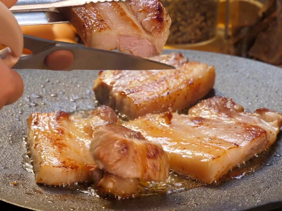 ジューシーに焼けた厚切り豚バラ肉を、熱い鉄板の上でキッチンバサミを使って小さめの短冊状にカットしている手元。