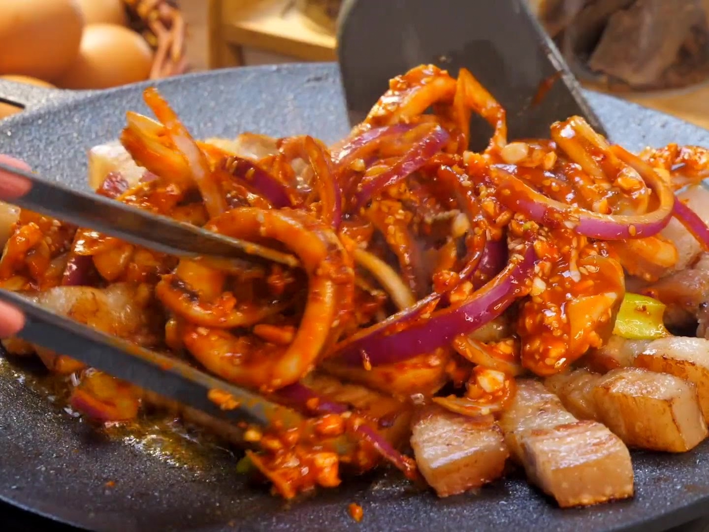 ジュージューと音を立てる鉄板の上で、スパチュラとトングを使って、味付けされたイカリング、豚バラ肉、赤玉ねぎを一緒に炒め合わせている様子。
