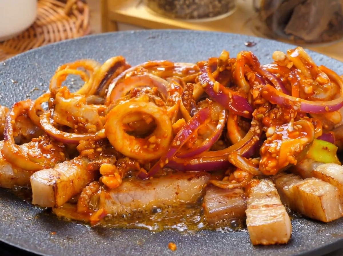 真っ赤なソースをまとった豚バラ肉とイカの炒め物がジュージューと泡立っている熱々の鉄板の上に、新鮮な青唐辛子のスライスがトッピングされている様子。