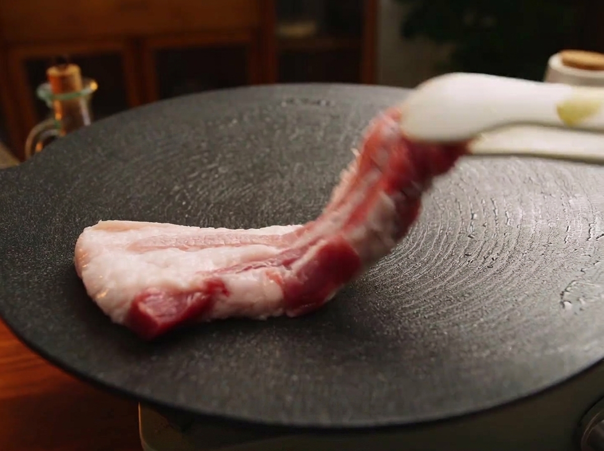 熱した黒い丸型の鉄板の上に、トングを使って厚切りの生の豚バラ肉を並べている様子。