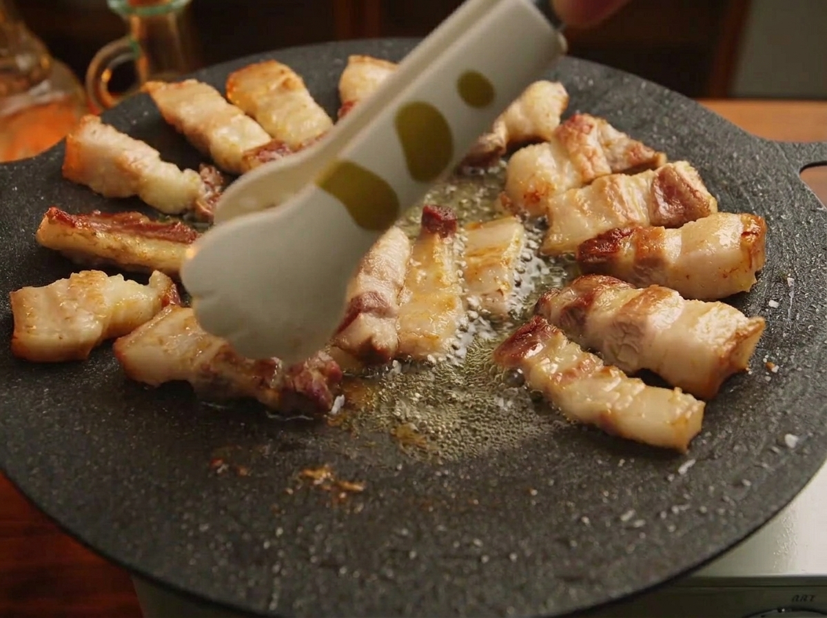 黒い鋳鉄製鉄板の縁に沿って、トングを使って一口大の焼き上がった豚バラ肉を円状に並べている様子。