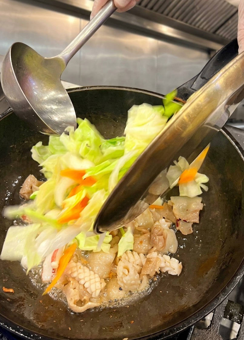 豚バラ肉とイカが入った熱い中華鍋に、ザク切りにしたキャベツや薄切りにした人参などの野菜を加えている様子。