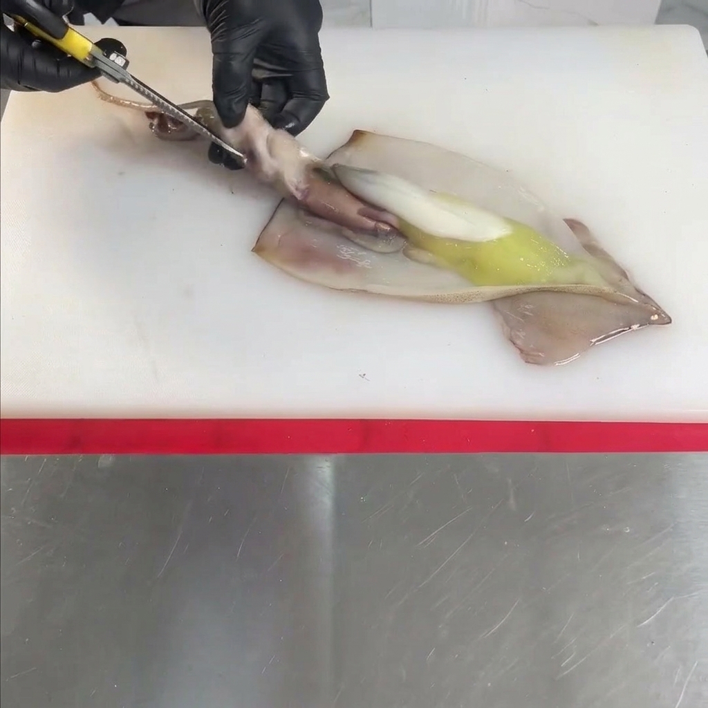 白いまな板の上で、手袋をはめた手がハサミを使って生のイカを切り開き、掃除している様子。