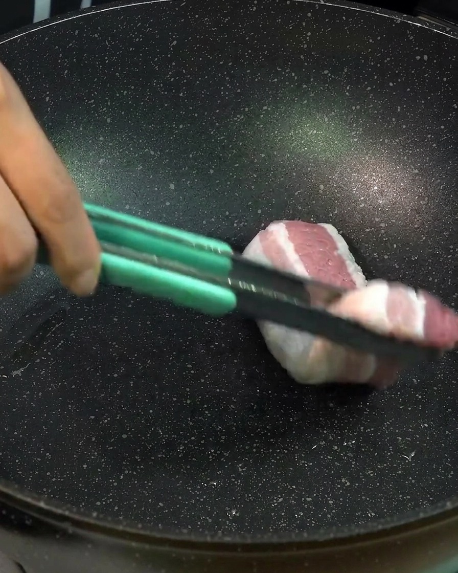 トングを使って、厚切りの生の豚バラ肉を黒いフライパンに並べて焼いている様子