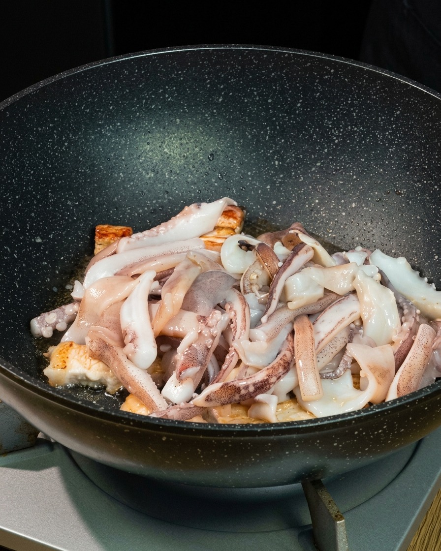 深さのある黒い中華鍋の中で、焼いた豚バラ肉の上に新鮮なイカをのせている様子