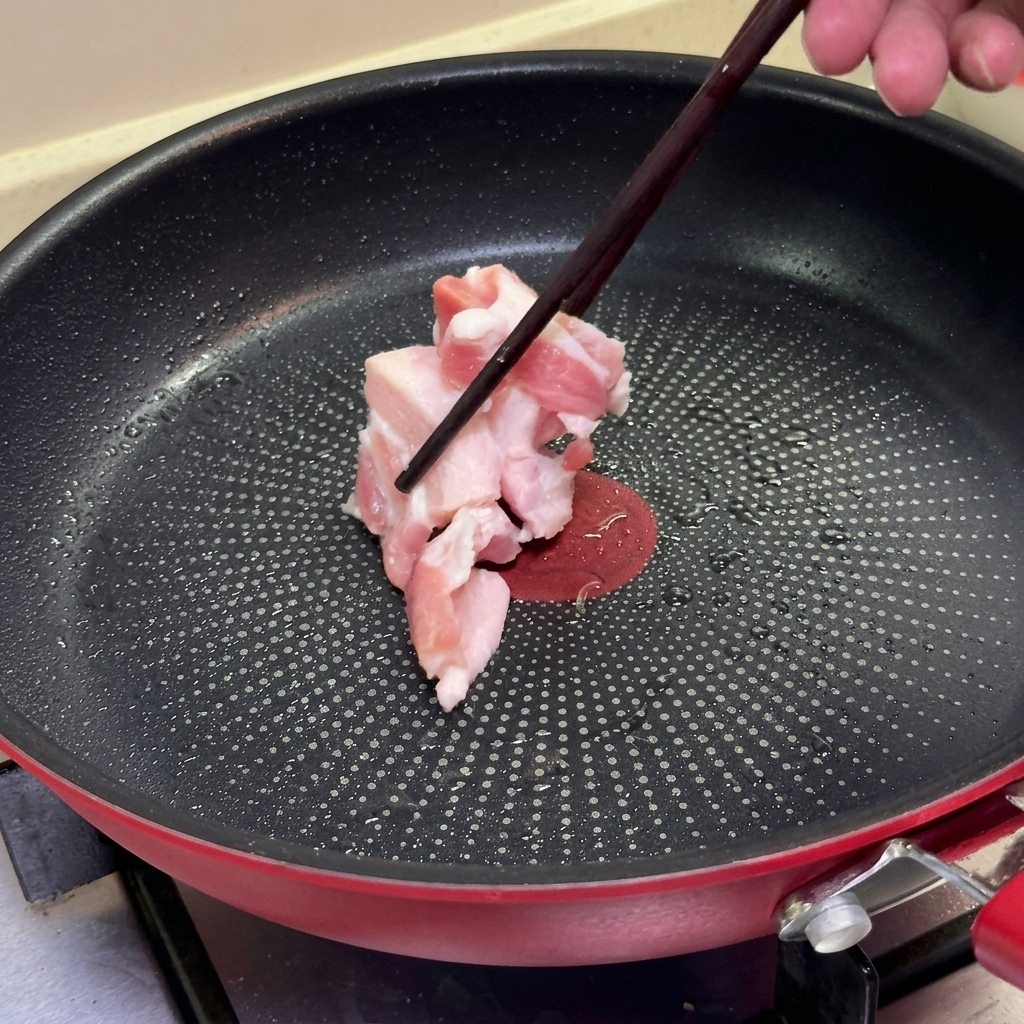 熱したフッ素樹脂加工의フライパンに、箸を使って生の豚バラ肉のスライスを並べている様子。