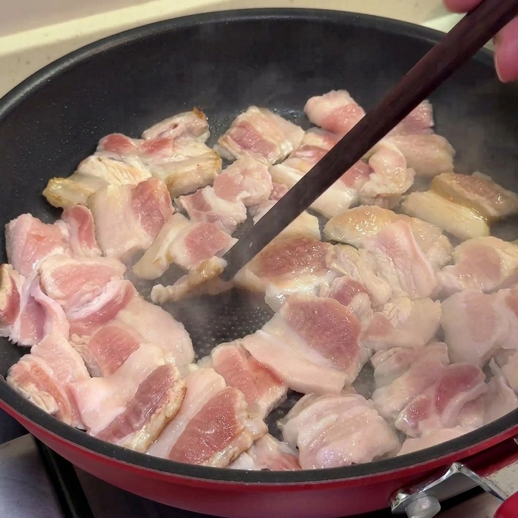 フッ素樹脂加工のフライパンで豚バラ肉を炒め、表面がきつね色に変わり、少し焦げ目がつくのを箸でひっくり返している様子。