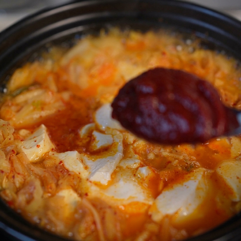 グツグツと沸騰するキムチと豆腐のスープの上で、スプーンに盛られた濃厚な深紅のコチュジャンを掲げている様子。