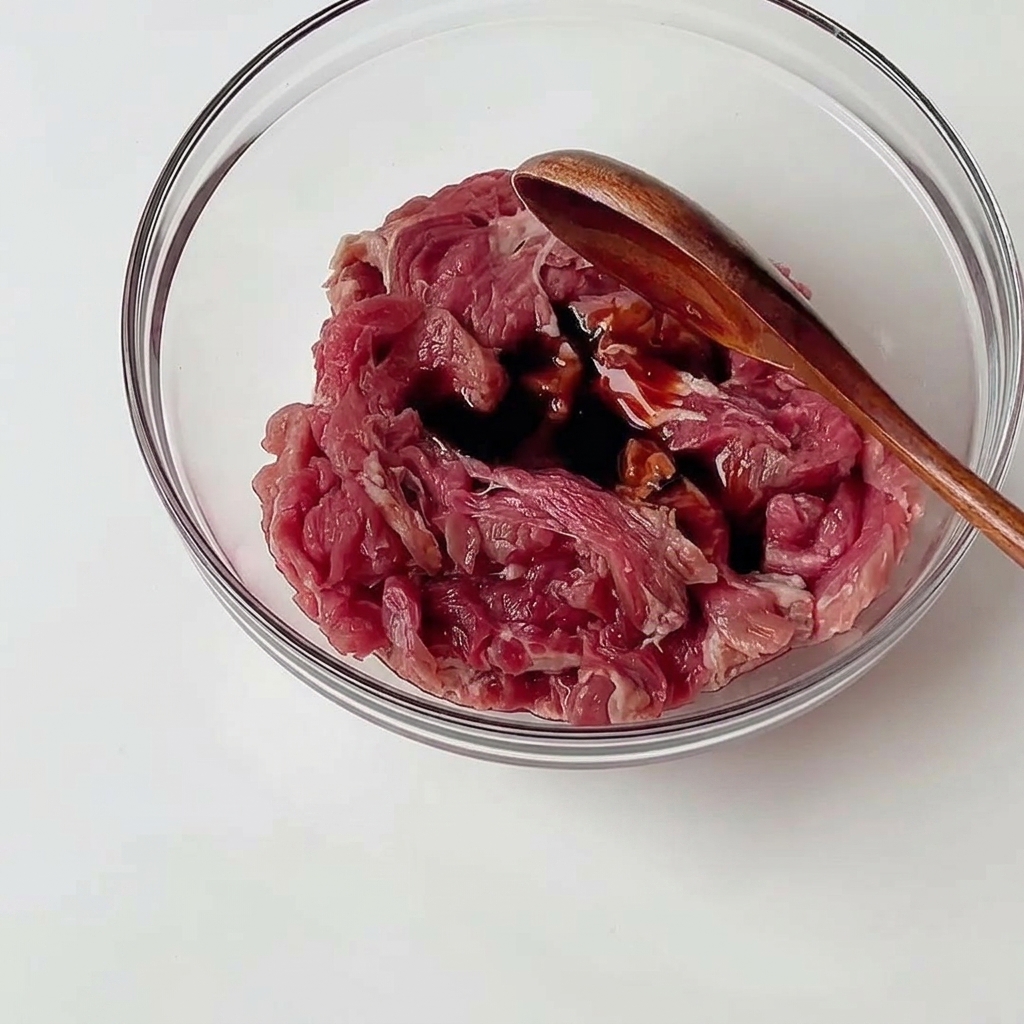 透明なガラスのボウルに入った牛薄切り肉に、木製のスプーンで濃い色の調味料を混ぜ合わせている様子。