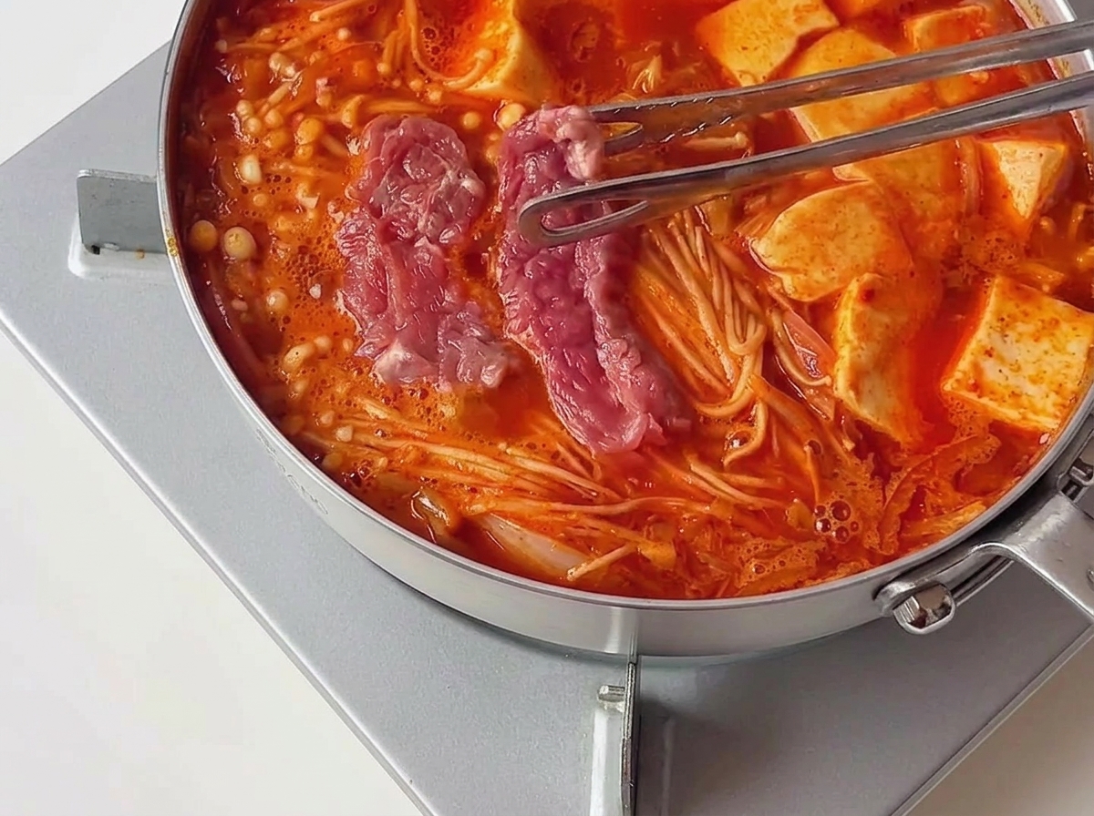 豆腐の横で熱々に沸騰している赤色のスープに、トングで下味のついたピンク色の生の牛肉を移し入れている様子。