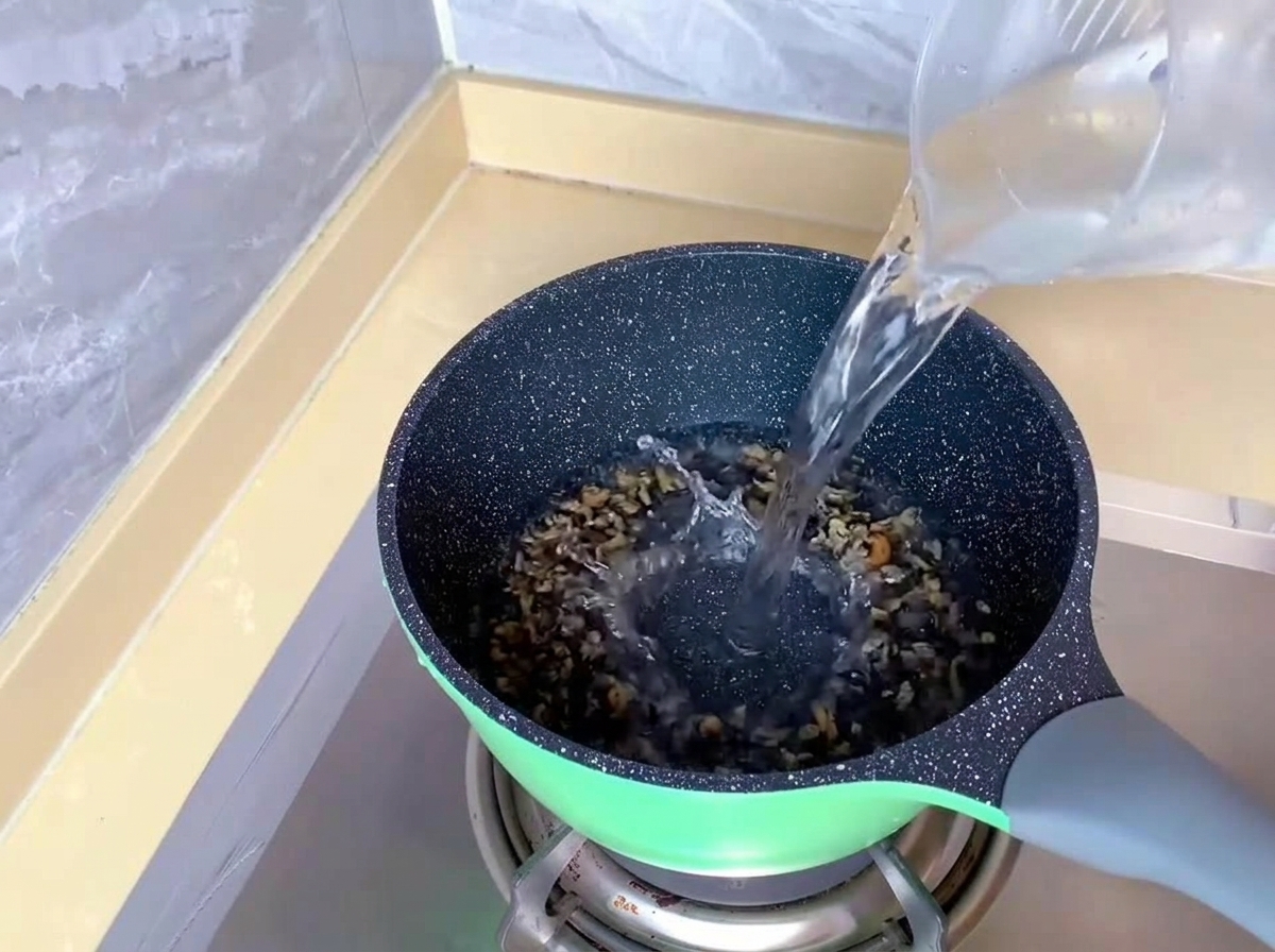 ガスコンロの上の緑色の鍋に、透明な計量カップから水が注がれている様子。