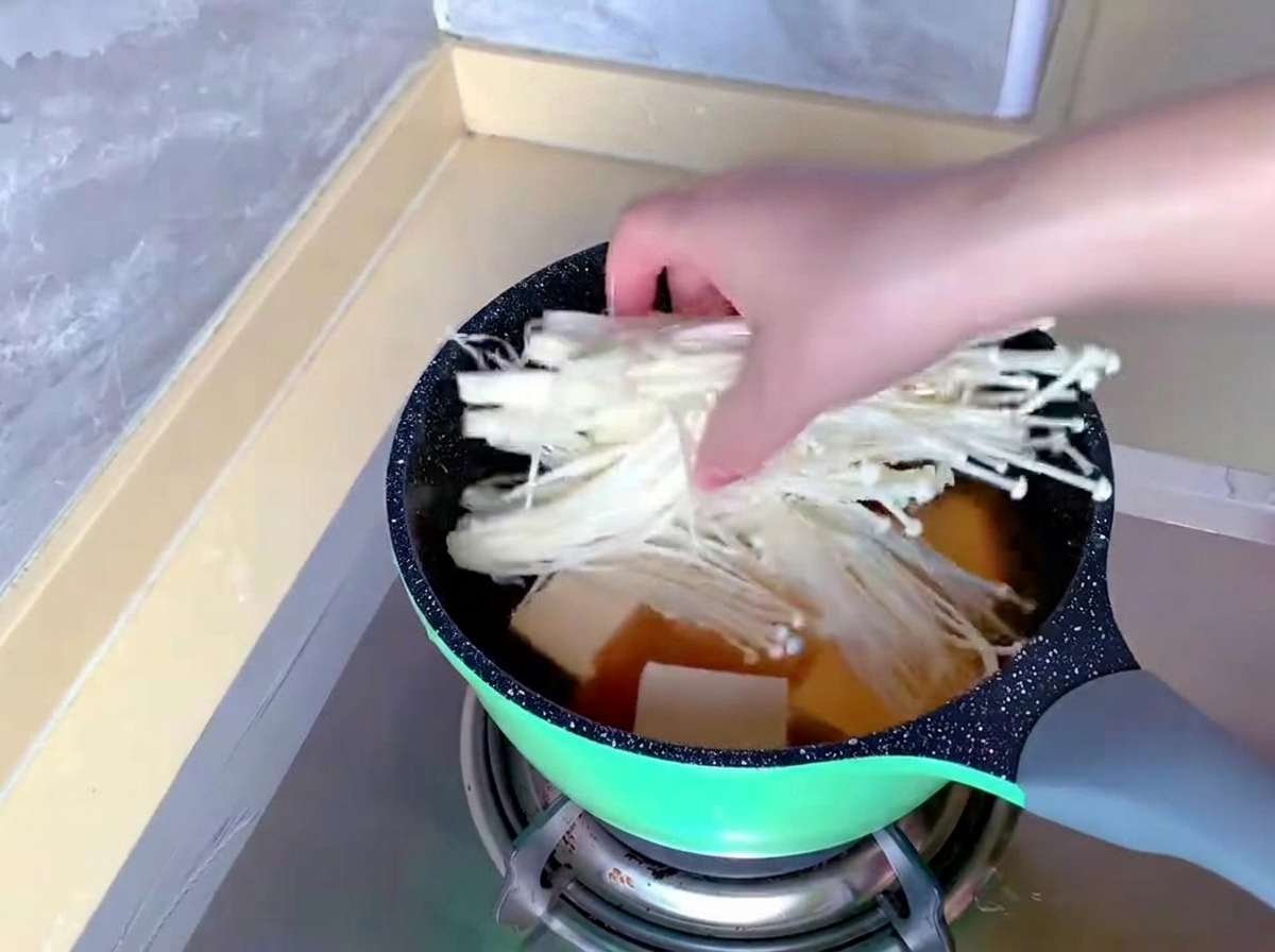 すでに豆腐とスープが入った鍋に、白くて長いえのき茸の束を手で入れている様子。