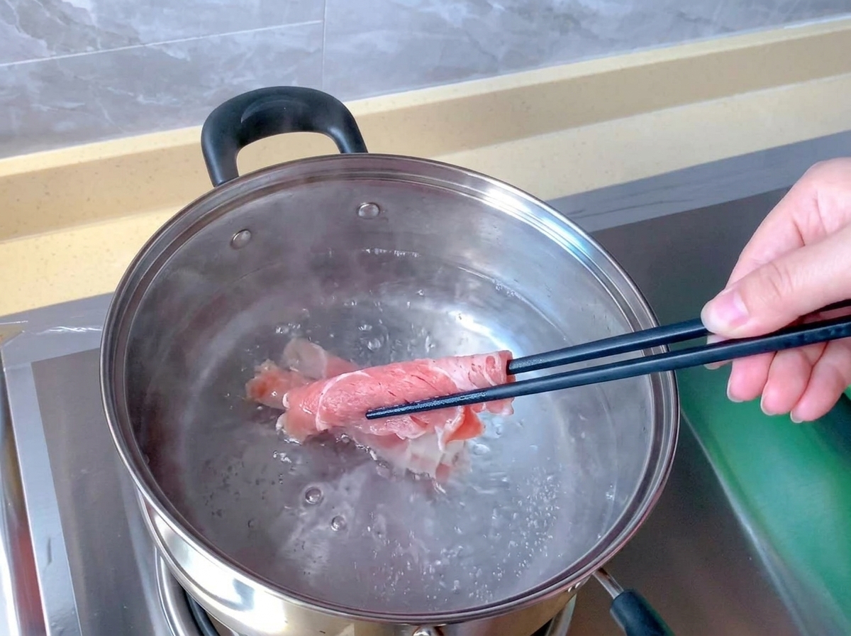 ステンレスの鍋で沸騰したお湯の中に、箸を使って生のピンク色の牛薄切り肉を浸している様子。