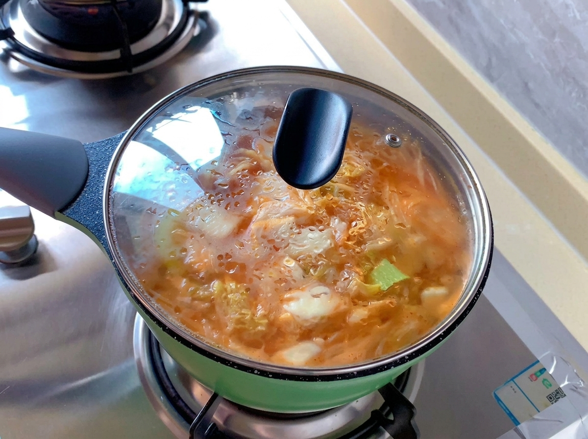 表面に水滴がついた透明なガラス蓋が閉められた、オレンジ色のキムチスープが入った緑色の小鍋。