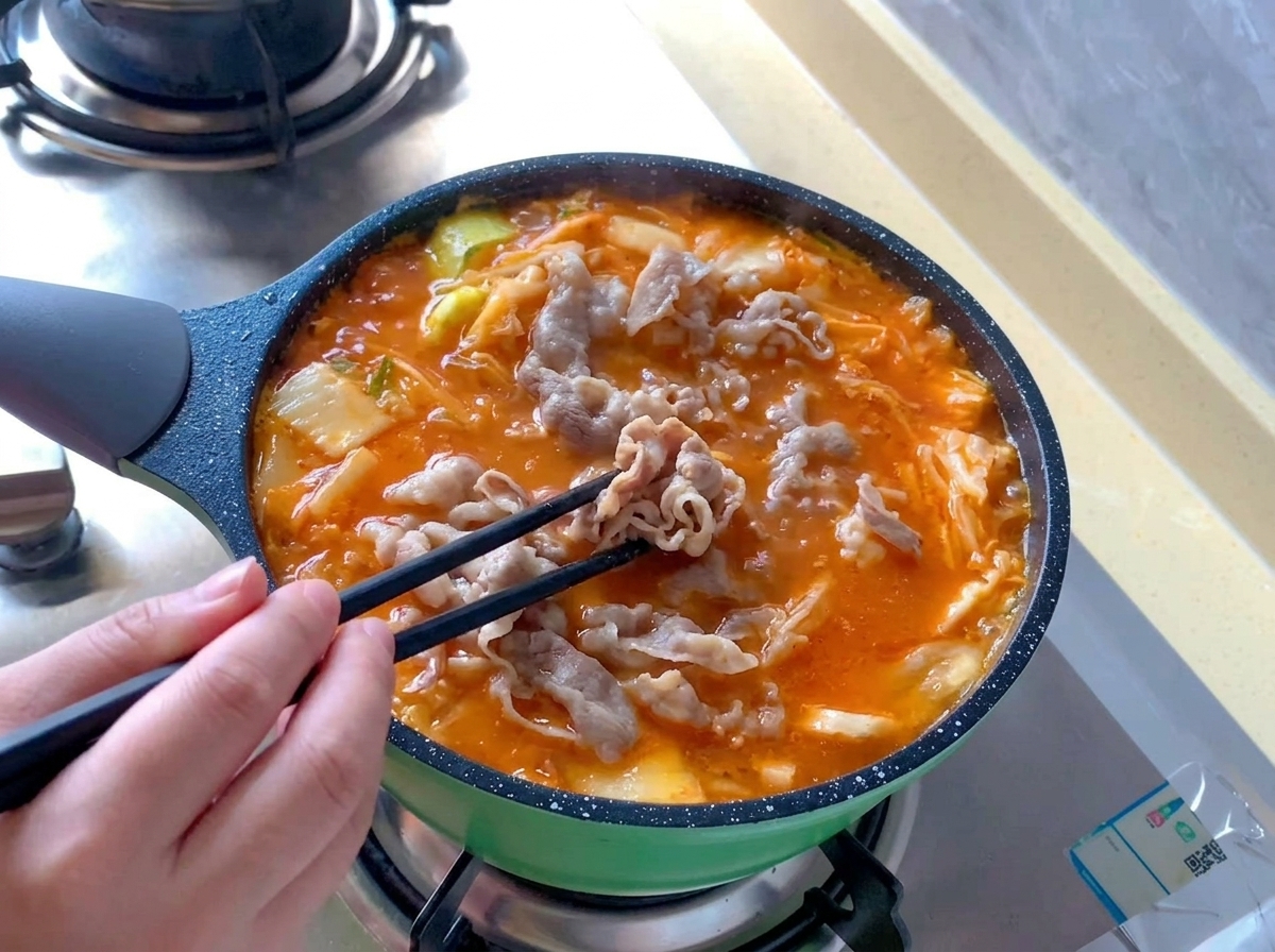 グツグツと煮立つ真っ赤なキムチスープの中に、黒い箸を使って湯通しした牛肉を広げながら入れている様子。